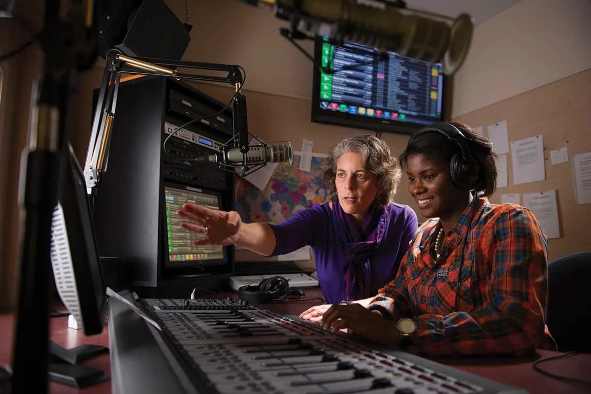 Student and instructor operating audio equipment in a radio broadcasting studio.